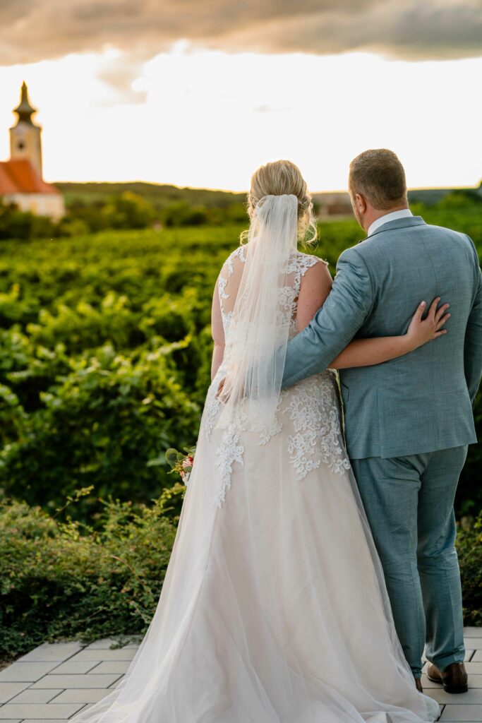 Brautpaar von hinten im Weinberg mit Kirchturm im Abendlicht – Sabrina Dötzl Fotografie Hollabrunn