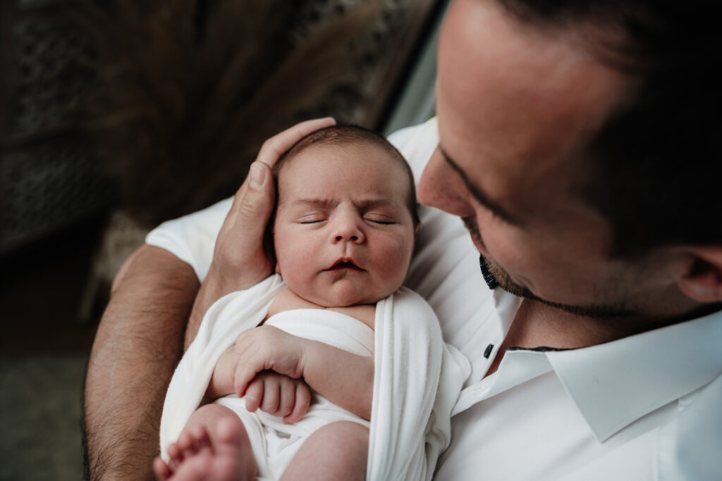 Papa hält sein Neugeborenes im Arm und blickt es liebevoll an – Sabrina Dötzl Fotografie Hollabrunn